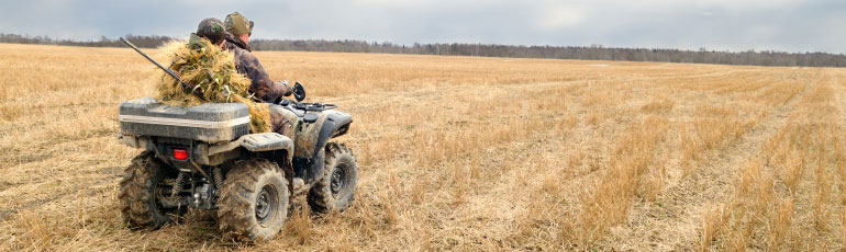 Two soldiers in camouflage riding an ATV in barren corn fields.